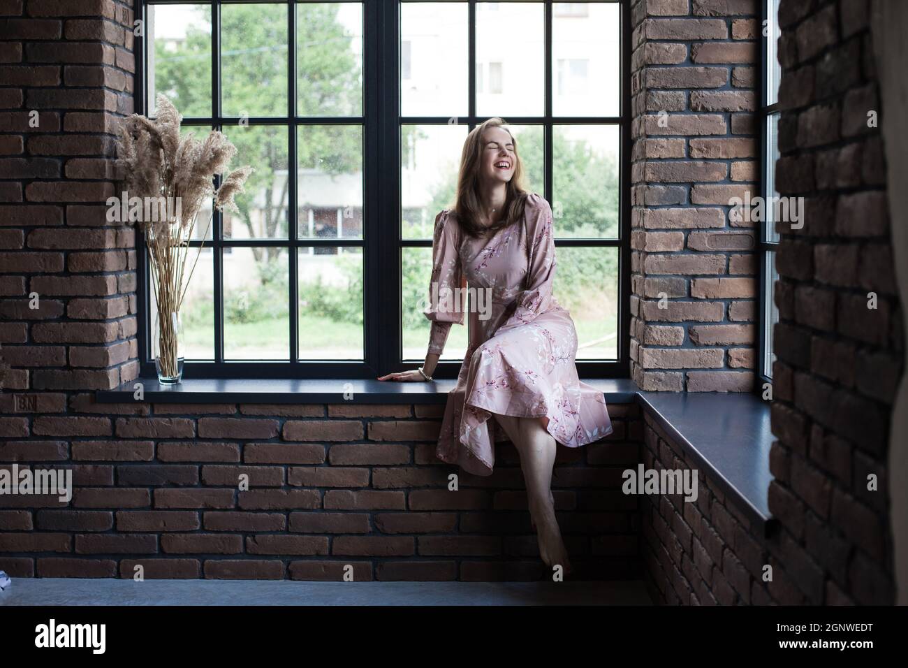 Beautiful young woman in pink dress sits on the windowsill barefoot ...