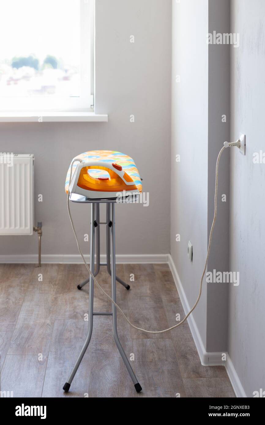 The electric iron is plugged into the socket on the ironing board in the  room against the background of the window. Iron clothes Stock Photo - Alamy