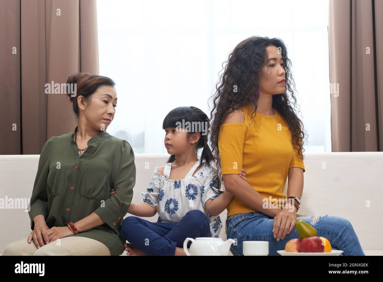 Little Asian girl sitting on sofa and trying to reconcile mother and grandmother being in quarrel Stock Photo