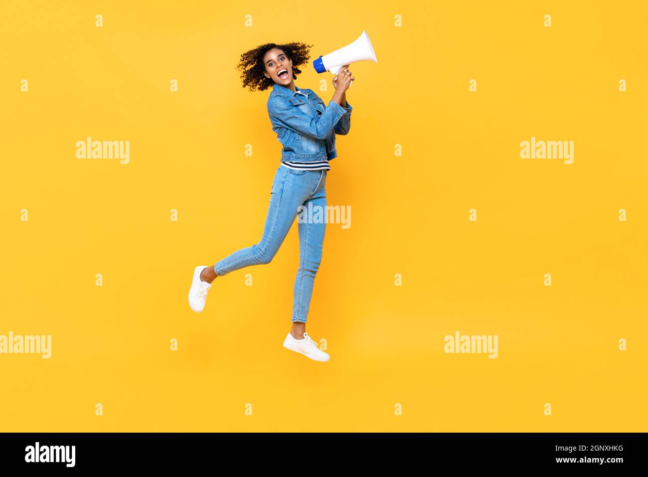 Cheerful young woman with megaphone jumping on yellow color studio background Stock Photo