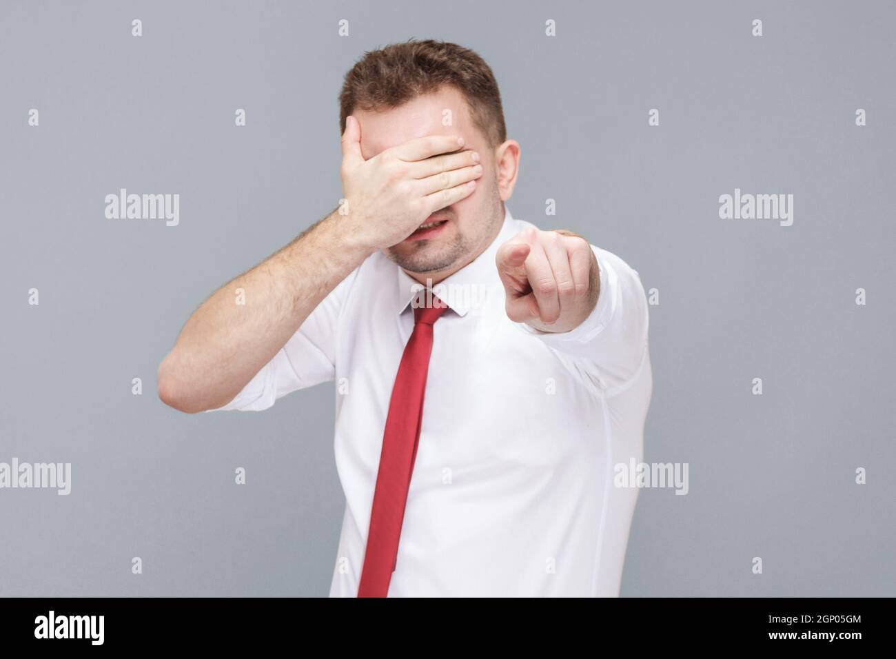 I don't want to look at you. Portrait of young angry shocked man in white shirt and tie standing and closed his eyes and pointing at camera and don't want to look. indoor isolated on gray background. Stock Photo