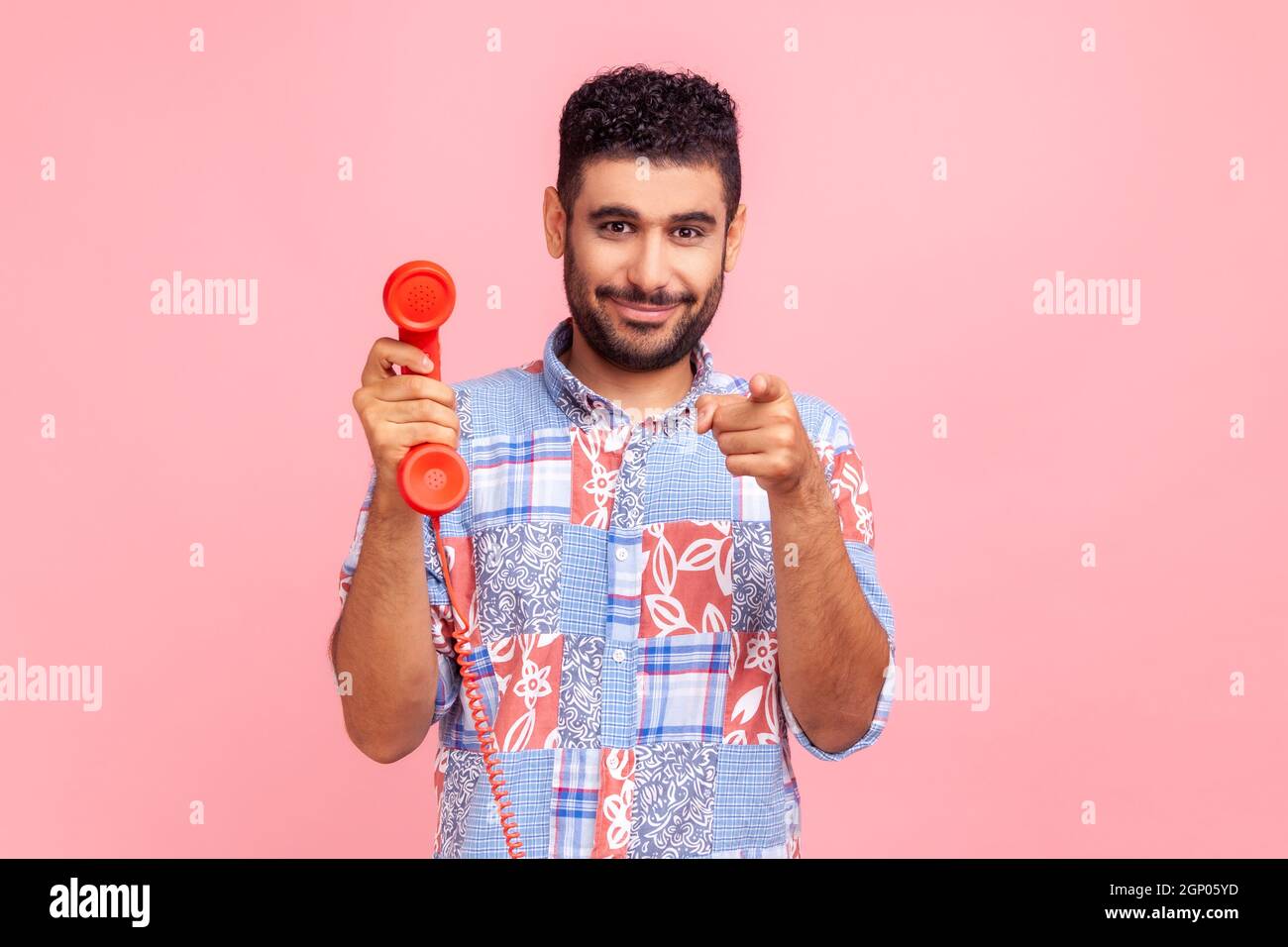 Smiling bearded man in blue casual shirt pointing finger on you, holding in hands red retro telephone, waiting for your call, looking at camera. Indoor studio shot isolated on pink background. Stock Photo