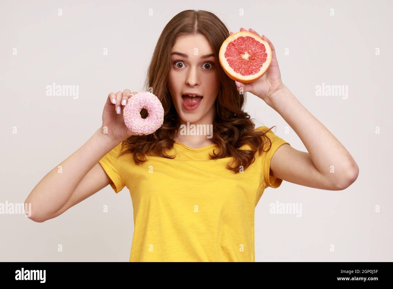 Playful excited teenager girl in yellow T-shirt holding half of ripe juicy grapefruit and round donut, looking camera with open mouth. Indoor studio shot isolated on gray background. Stock Photo