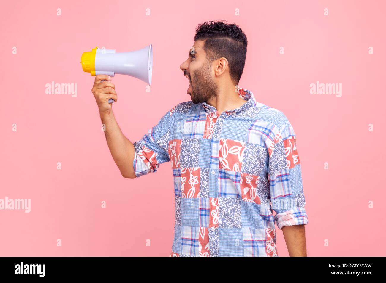 Profile of young adult excited crazy bearded man wearing blue casual style shirt screaming holding loudspeaker megaphone, looking aside at loud speaker. Indoor studio shot isolated on pink background. Stock Photo