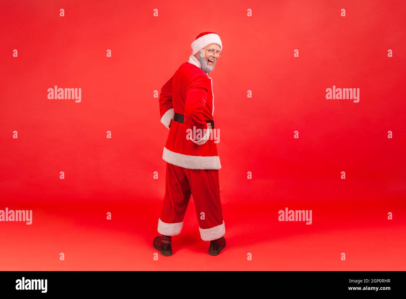 Full length portrait of elderly man with gray beard in santa claus costume standing backwards with hands on hips, turning to camera with excited look. Indoor studio shot isolated on red background. Stock Photo