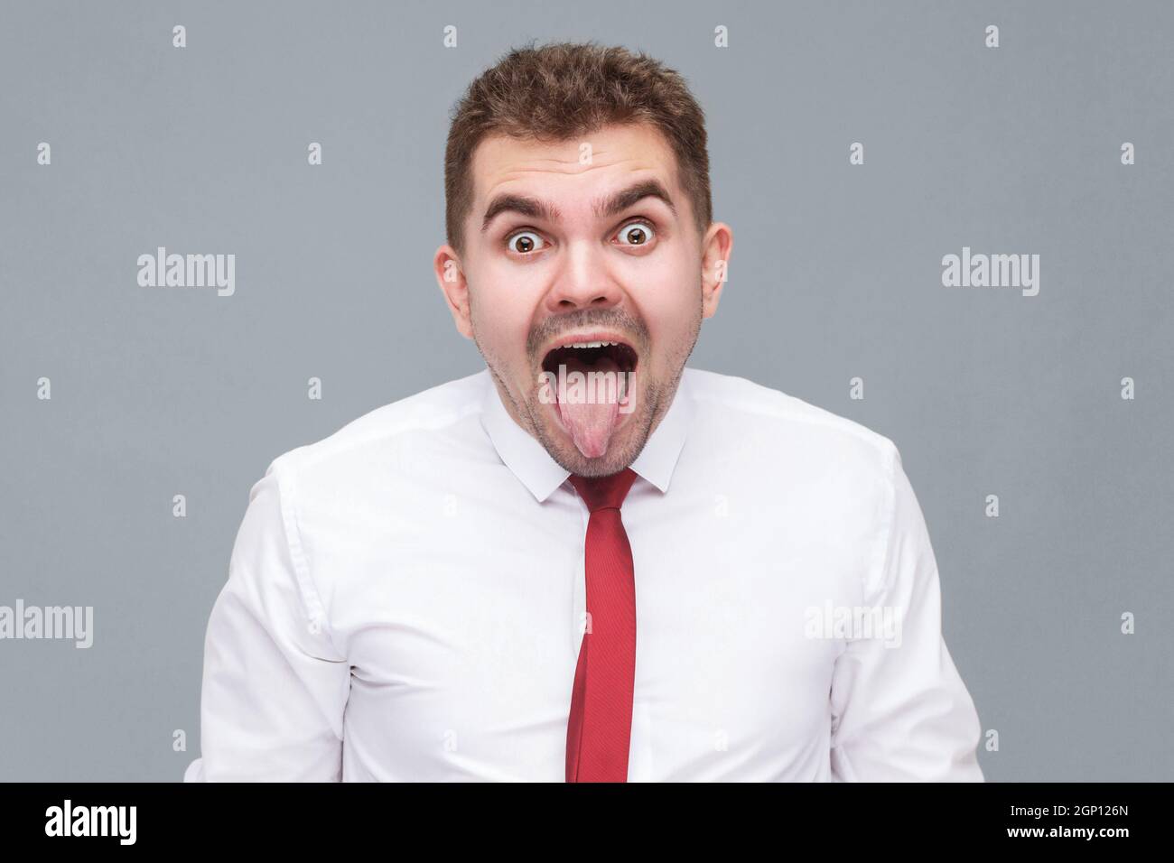 Portrait of young funny crazy man in white shirt and tie standing with big eyes and tounge out and looking at camera with crazy funny face. indoor isolated on gray background. Stock Photo