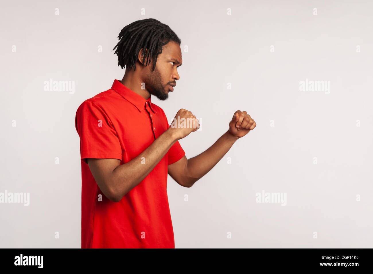 Profile portrait angry of man with dreadlocks wearing red casual style T-shirt, punching, boxing with clenched fists, being ready fighting. Indoor studio shot isolated on gray background. Stock Photo