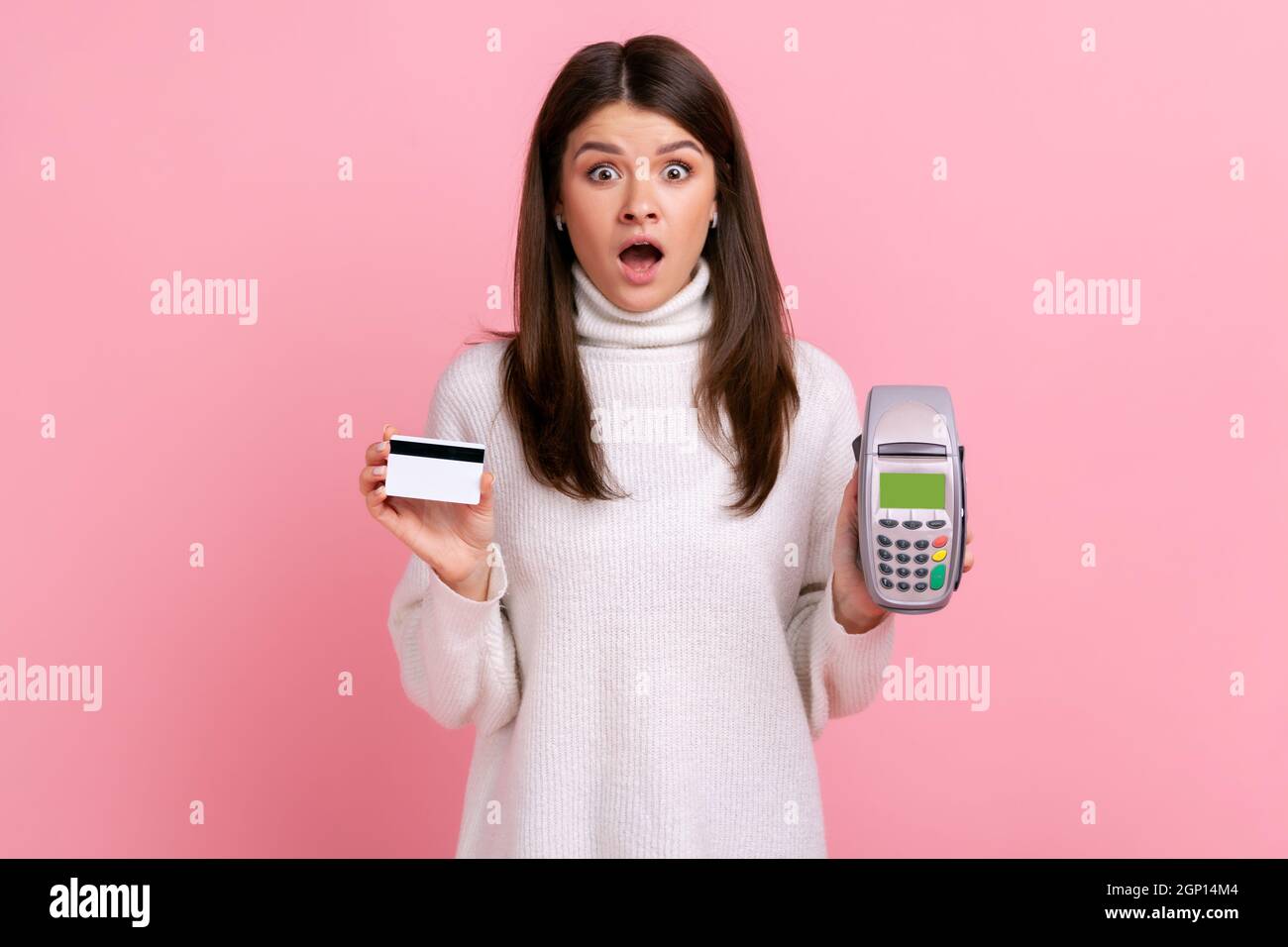 Astonished woman showing pos payment terminal and credit or debit card, using cashless payments, nfc, wearing white casual style sweater. Indoor studio shot isolated on pink background. Stock Photo