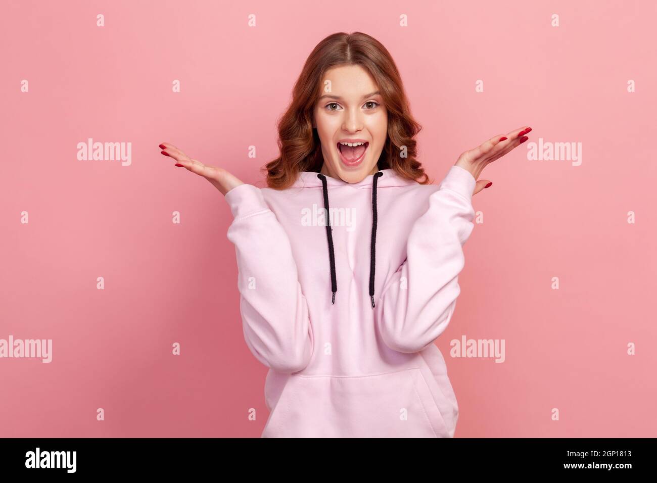 Portrait of extremely surprised teen girl in hoodie looking at camera with big eyes and open mouth, shocked with sudden victory. Indoor studio shot, isolated on pink background Stock Photo