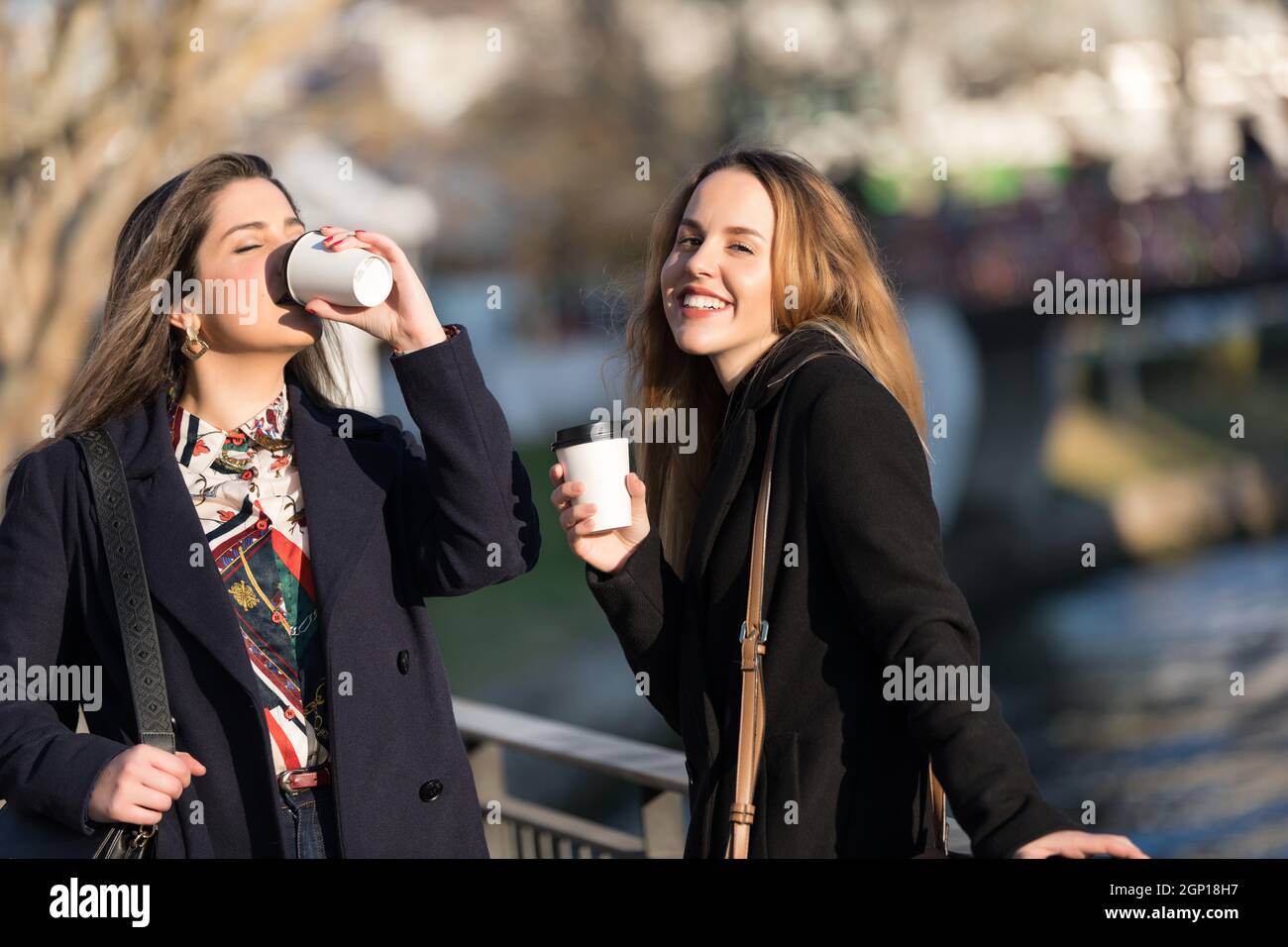 Outdoors fashion portrait of two young beautiful women friends drinking coffee. Smiling and going shopping. Kissing a cup of coffee. Bright make up Stock Photo