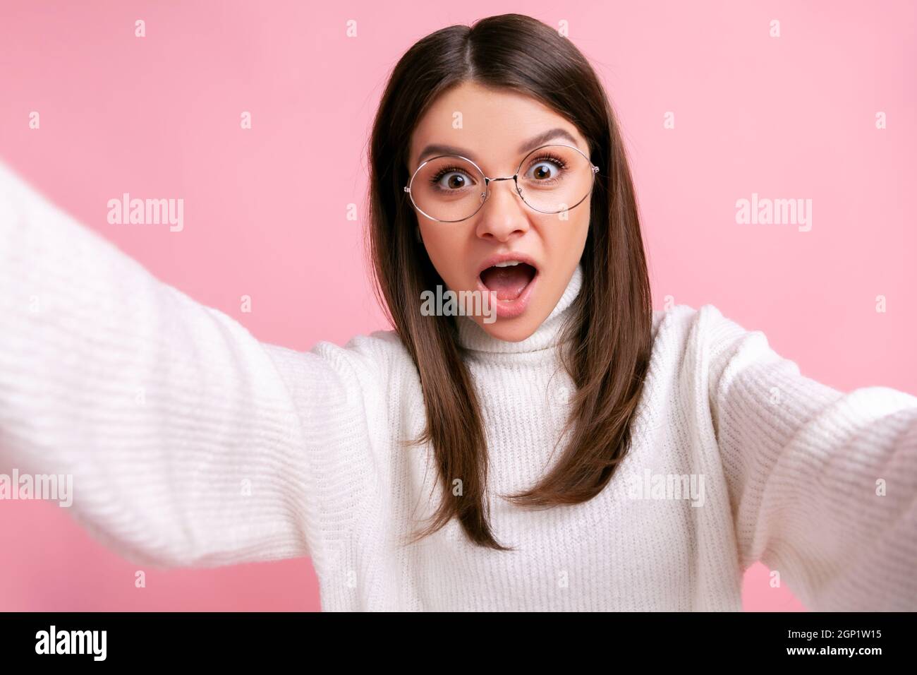 Shocked young adult female taking selfie, having astonished and scared look, point of view photo, wearing white casual style sweater. Indoor studio shot isolated on pink background. Stock Photo