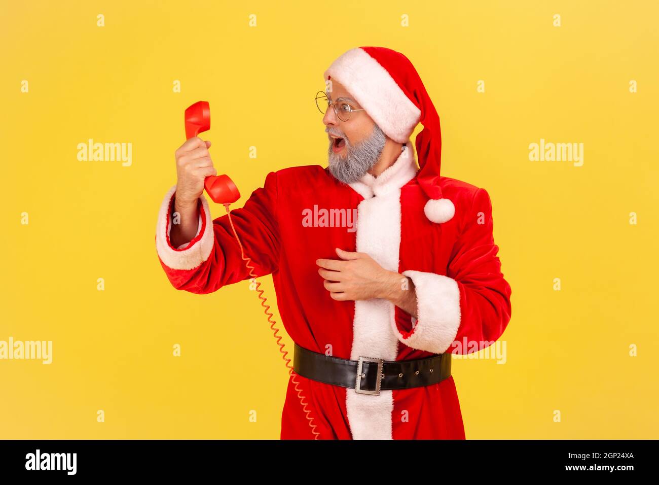 Side view of elderly man with gray beard wearing santa claus costume holding retro handset, looking at land line phone with open mouth. Indoor studio shot isolated on yellow background. Stock Photo