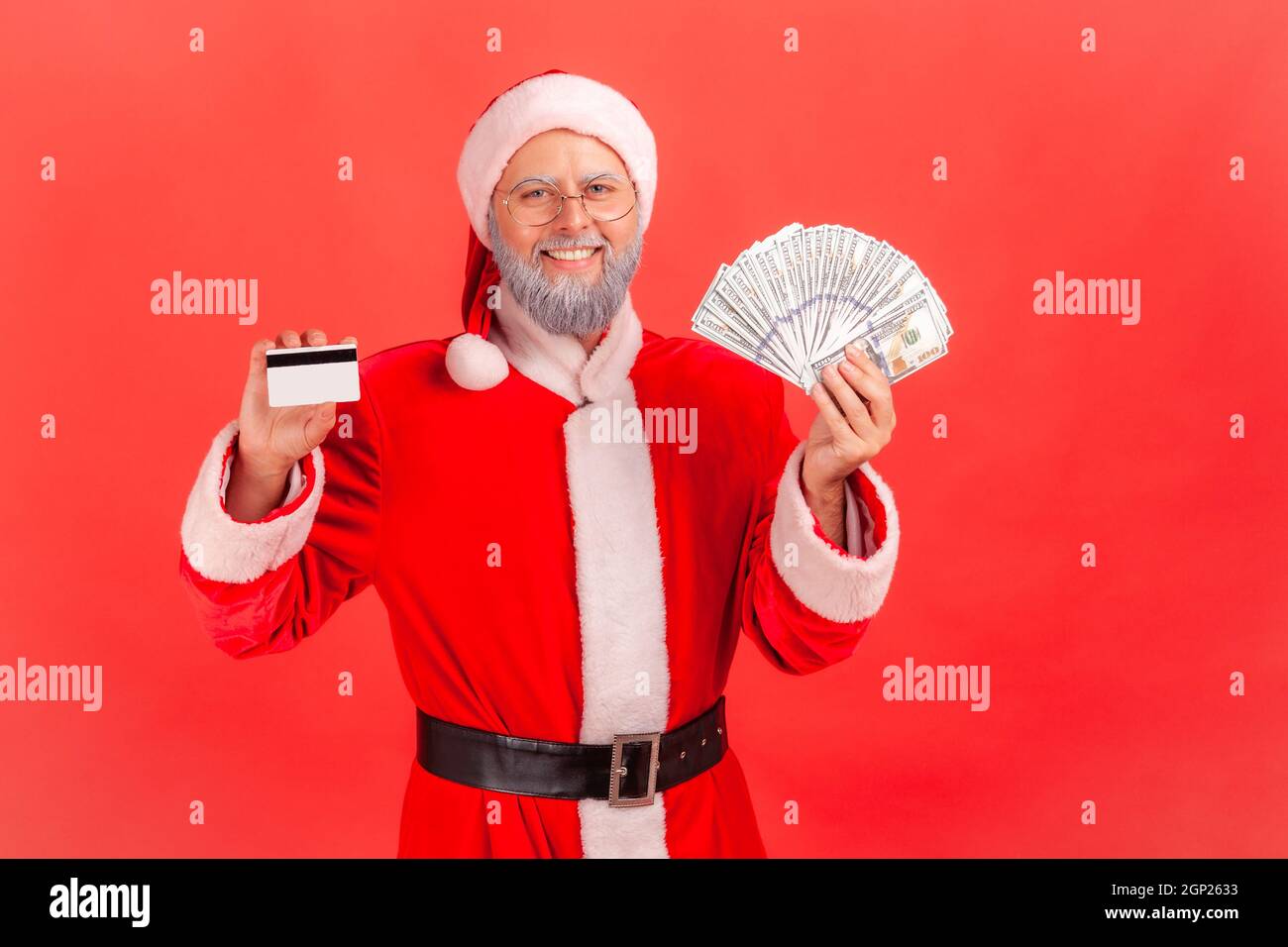 Satisfied elderly man with gray beard wearing santa claus costume standing holding fan of dollars and showing credit card, cashback. Indoor studio shot isolated on red background. Stock Photo