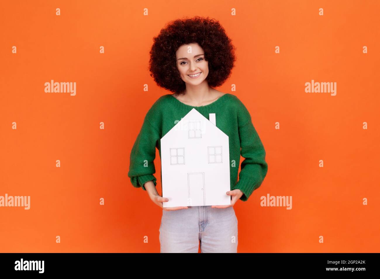 Pleasant looking woman with Afro hairstyle wearing green casual style sweater holding white paper house, buying new apartment, real estate agency. Indoor studio shot isolated on orange background. Stock Photo
