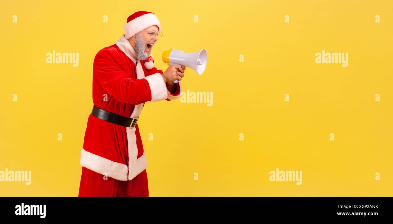 Side view of angry screaming elderly man with gray beard wearing santa claus costume holding megaphone and yelling with aggressive expression. Indoor studio shot isolated on yellow background. Stock Photo