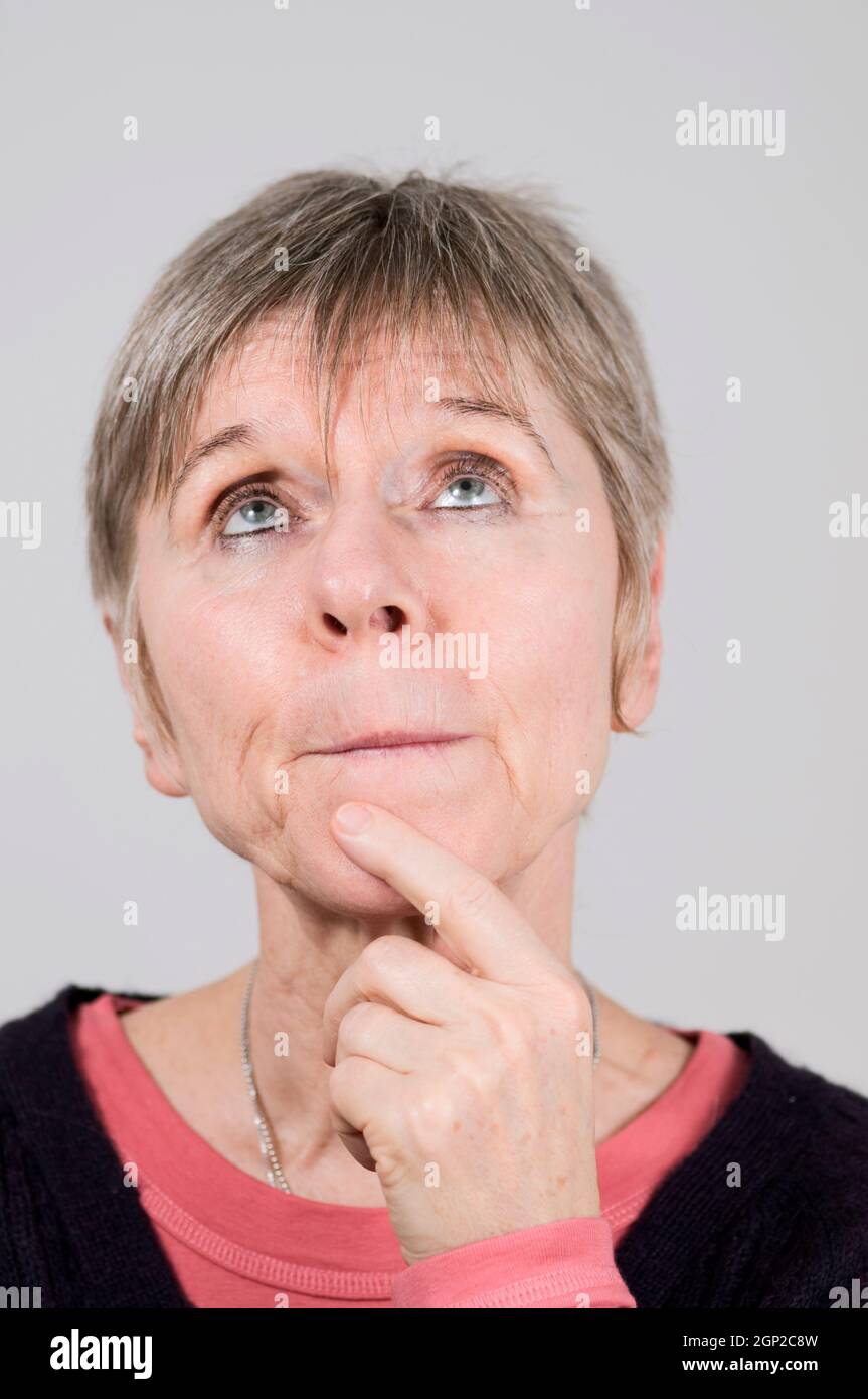 Head portrait of an older woman in front of a white background pensive and pressing lips, looking up with the left index finger on her chin. Stock Photo
