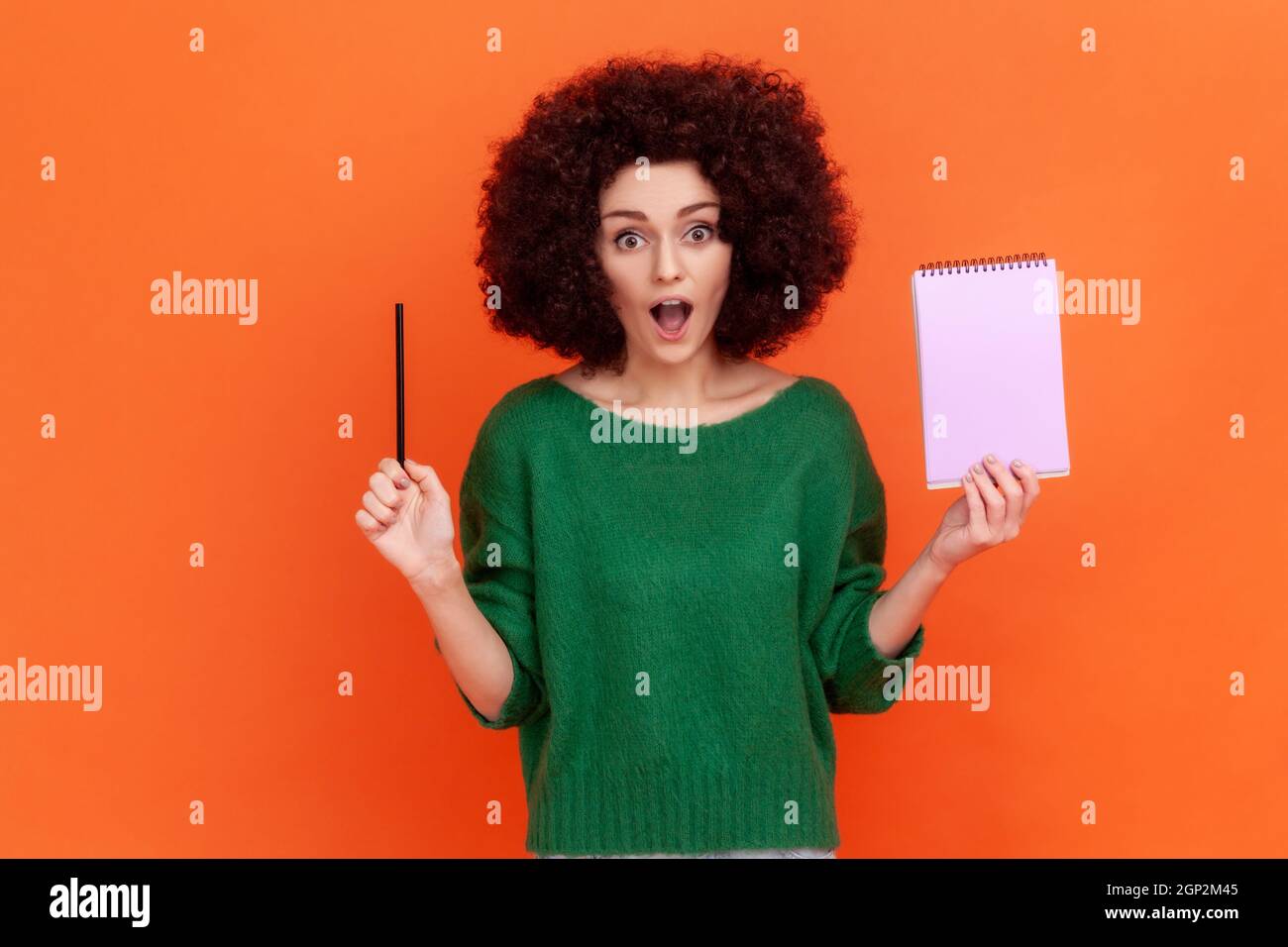 Surprised woman with Afro hairstyle wearing green casual style sweater standing with open mouth, holding paper notebook and pencil. Indoor studio shot isolated on orange background. Stock Photo