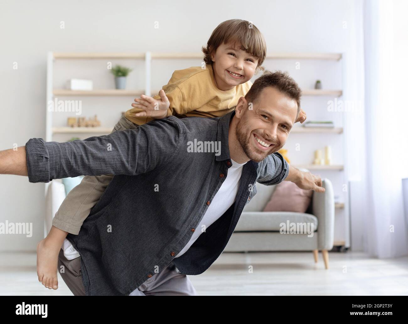 Happy time at home. Positive young father piggybacking his little son and smiling to camera, playing plane game together at living room Stock Photo
