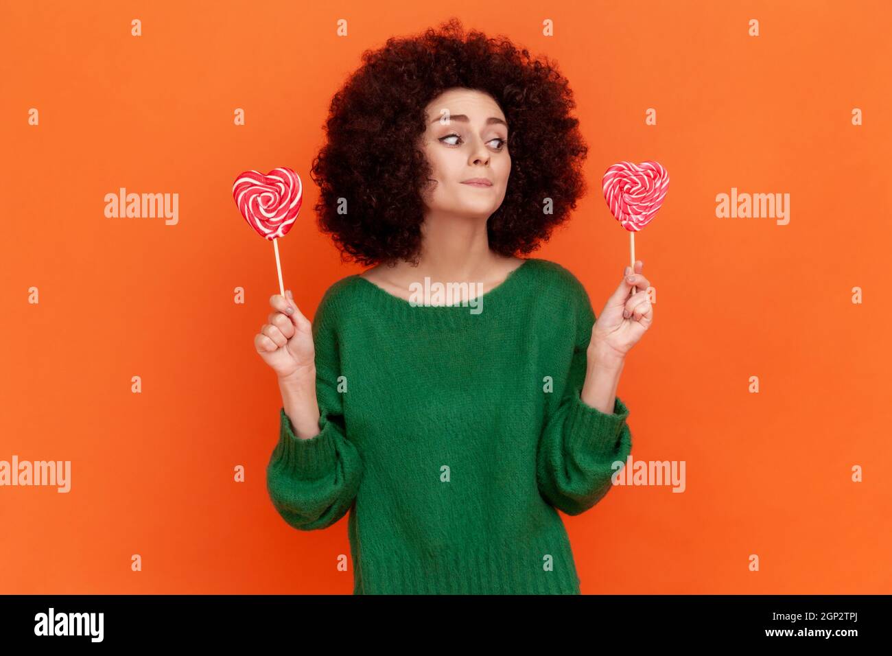 Attractive woman with Afro hairstyle wearing green casual style sweater keeping diet, decides eat candy or not, looking at sweets. Indoor studio shot isolated on orange background. Stock Photo
