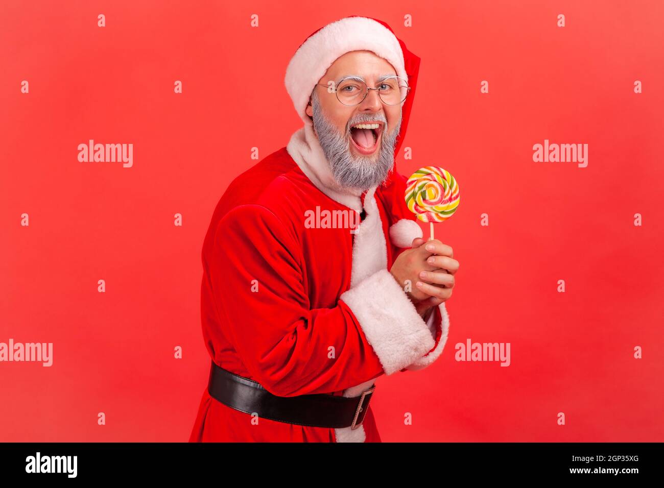 Portrait of happy amazed smiling elderly man with gray beard wearing santa claus costume standing with sugary lollypop in hands, looking at camera. Indoor studio shot isolated on red background. Stock Photo