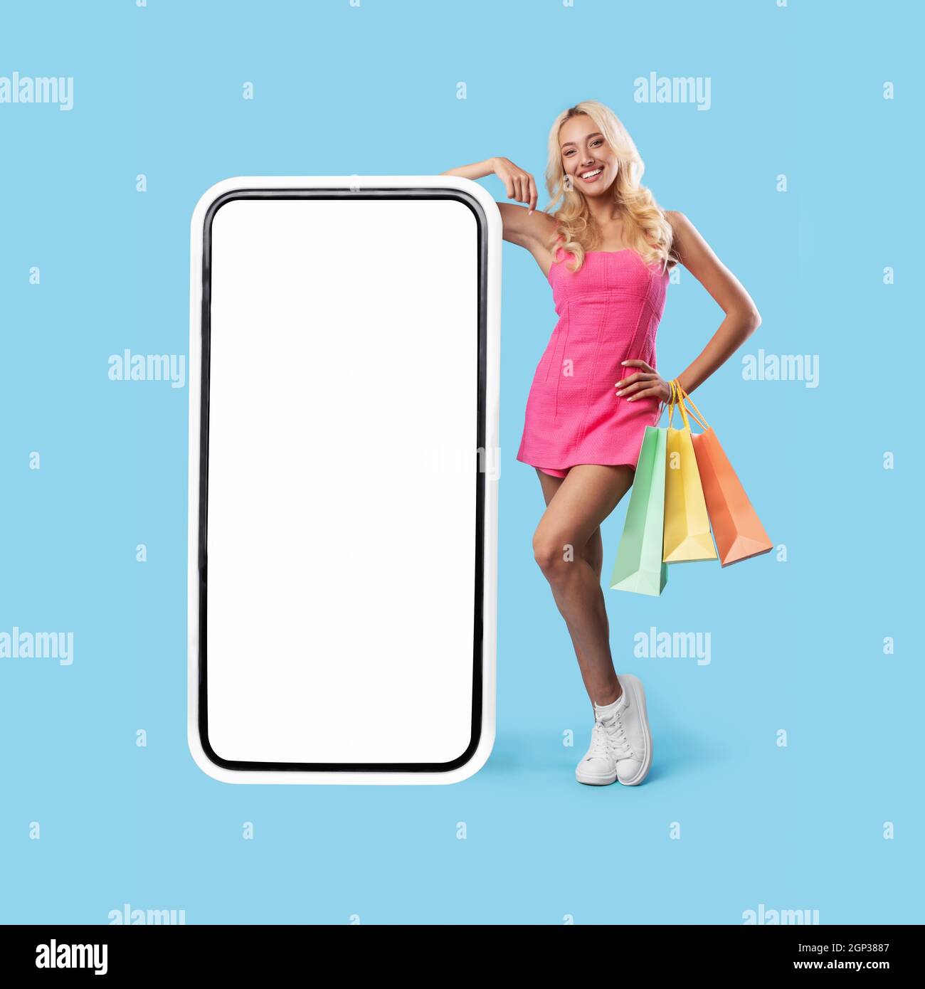 Mobile Offer. Happy pretty blonde lady leaning on huge cellphone with empty white screen, holding shopper bags, recommending new app or webshop Stock Photo - Alamy