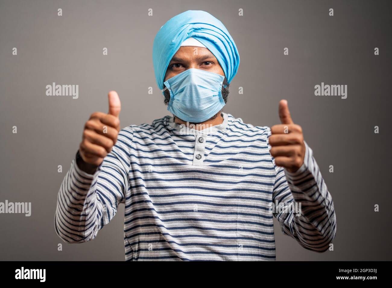 Young sikh man with medical face mask recommending to wear face mask by showing thumbs up gesture during coronavirus covid-19 pandemic. Stock Photo