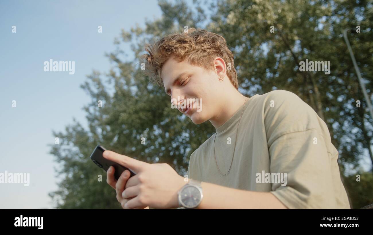 Handsome blond hair colored eyed boy while surfing the internet getting new message notification and answers with a smile. Beautiful nature and trees Stock Photo