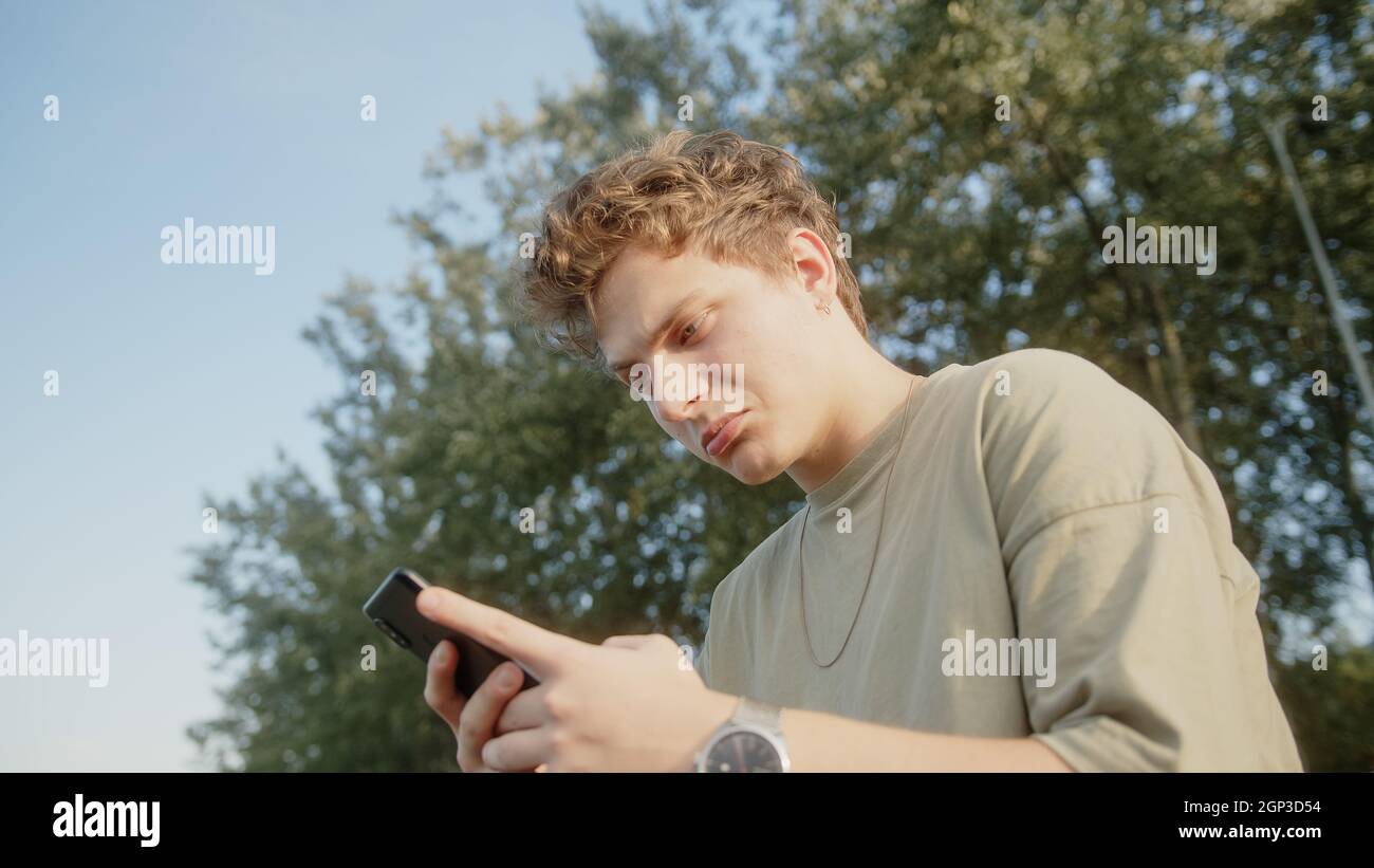 Handsome blond hair colored eyed boy while surfing the internet getting new message notification and answers with a smile. Beautiful nature and trees Stock Photo