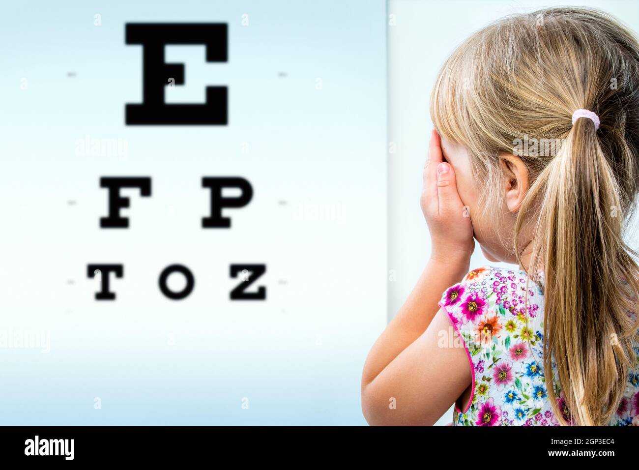 Close up rear view of girl testing eyesight. Infant closing one eye with hand looking at vision test chart. Stock Photo