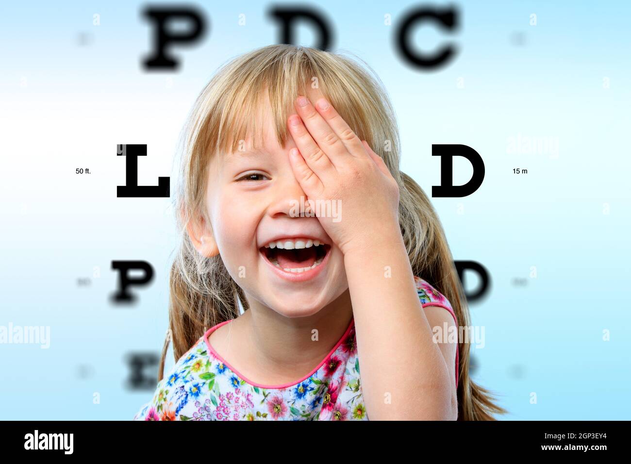 Close up face portrait of happy girl having fun at vision test.Conceptual image with girl closing one eye with hand and block letter eye chart in back Stock Photo