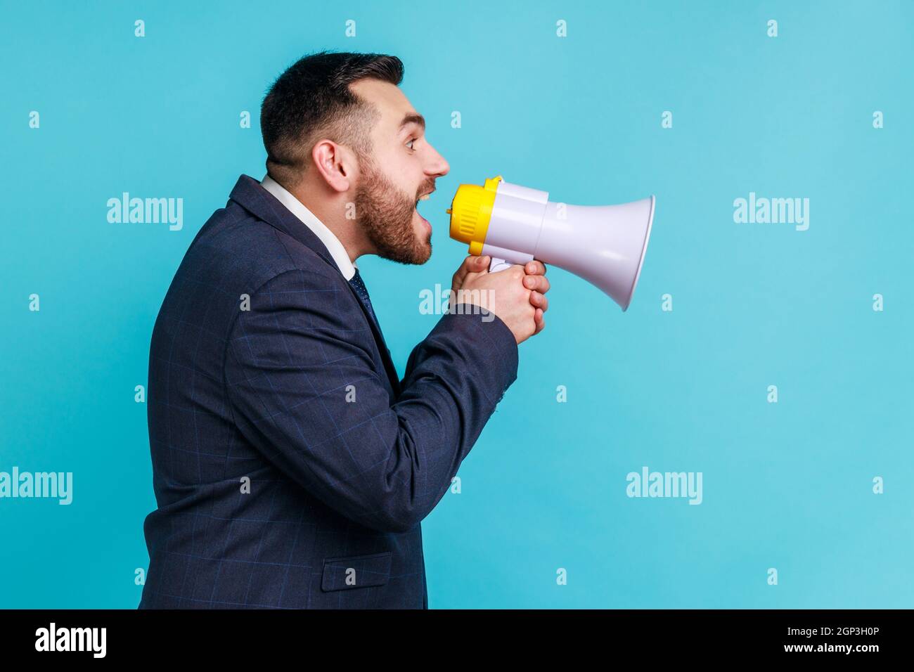 Side view portrait of angry bossy wearing official style suit loudly screaming at bullhorn megaphone holding hand on forehead and looking at distance. Indoor studio shot isolated on blue background. Stock Photo