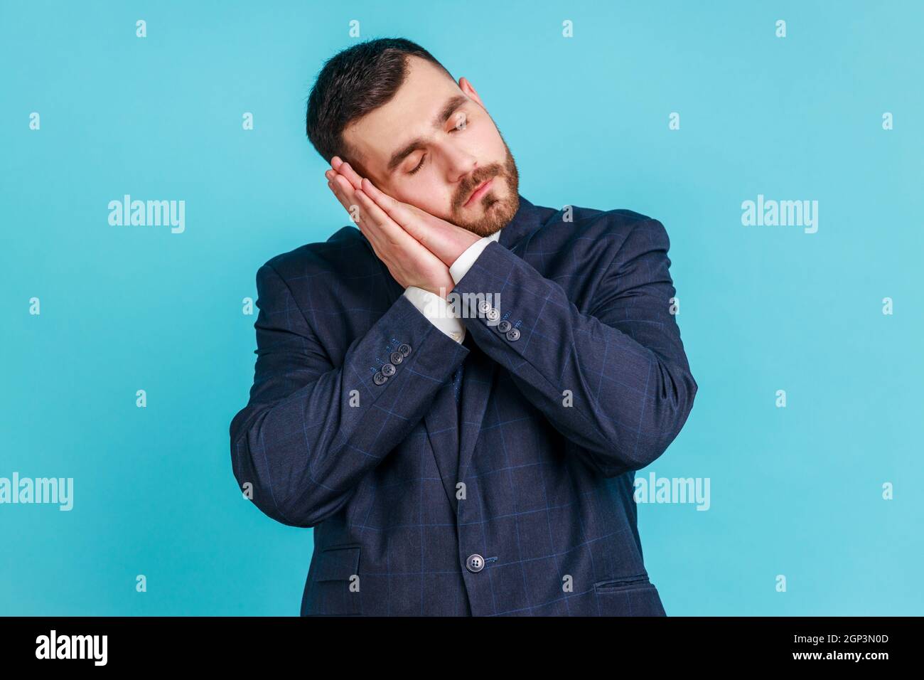 Bedtime. Portrait of handsome bearded man in suit sleeping laying down on her palms and smiling pleased, having comfortable nap and resting, dozing off. Indoor studio shot isolated on blue background. Stock Photo