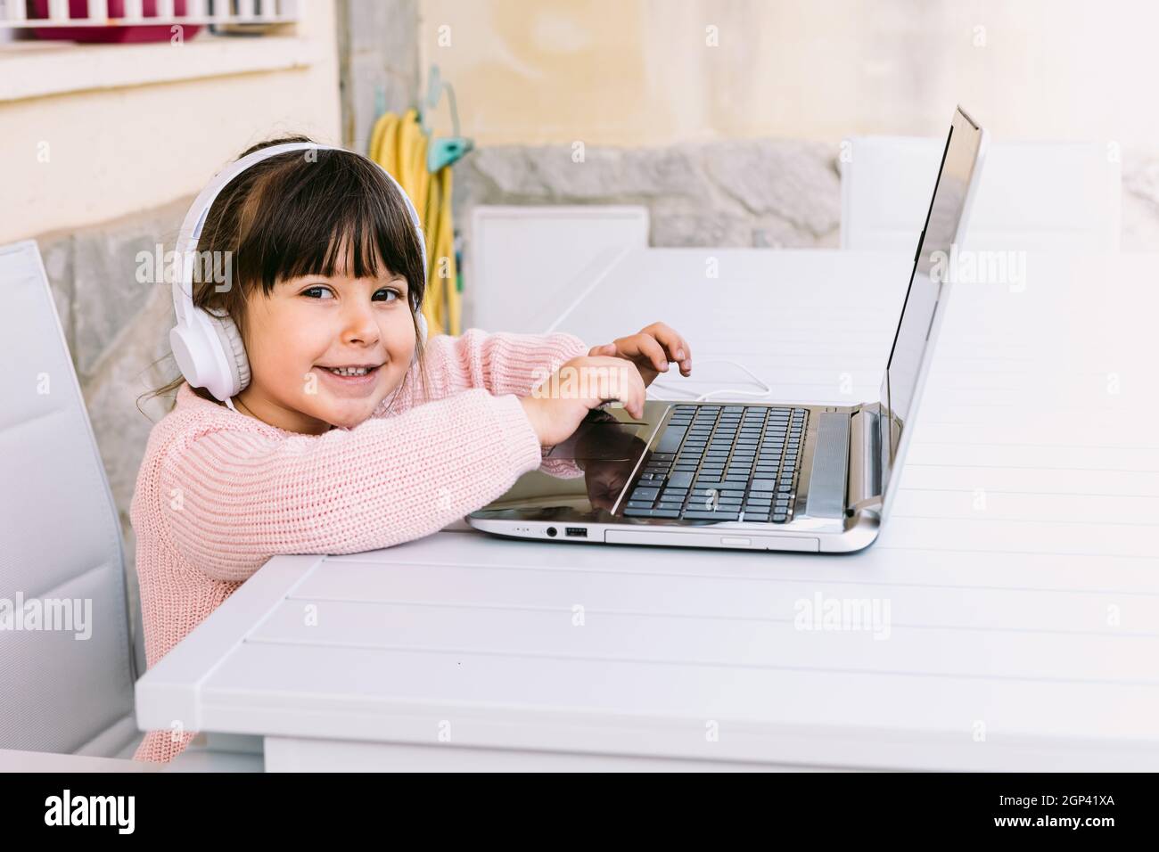 Little girl with white headphones, wearing pink sweater, sitting in front of laptop, typing, on terrace, smiling. Concept of distance studying online, Stock Photo