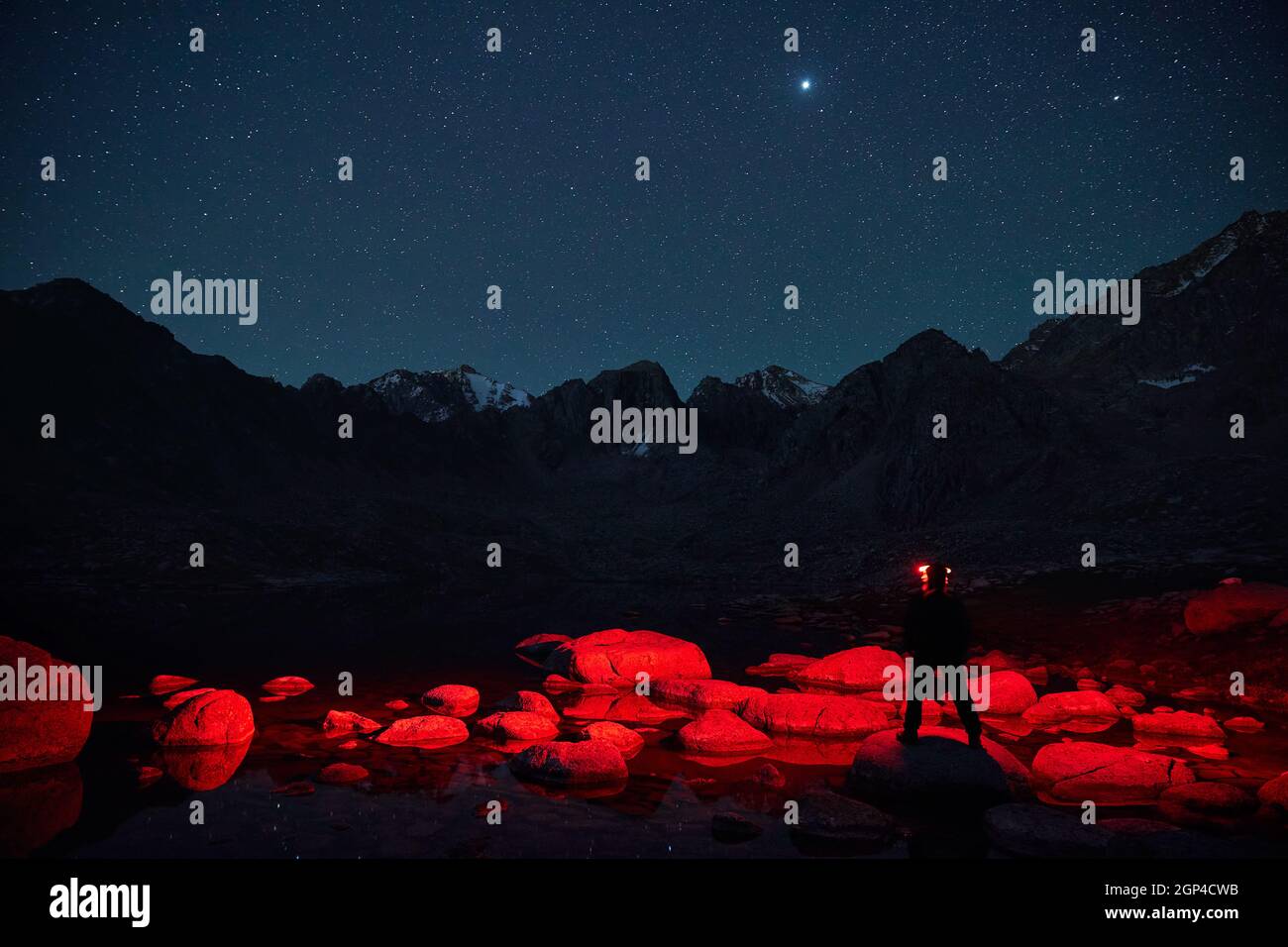 Tourist man in silhouette with red light of headlamp near lake in the mountains under night sky with stars. Stock Photo
