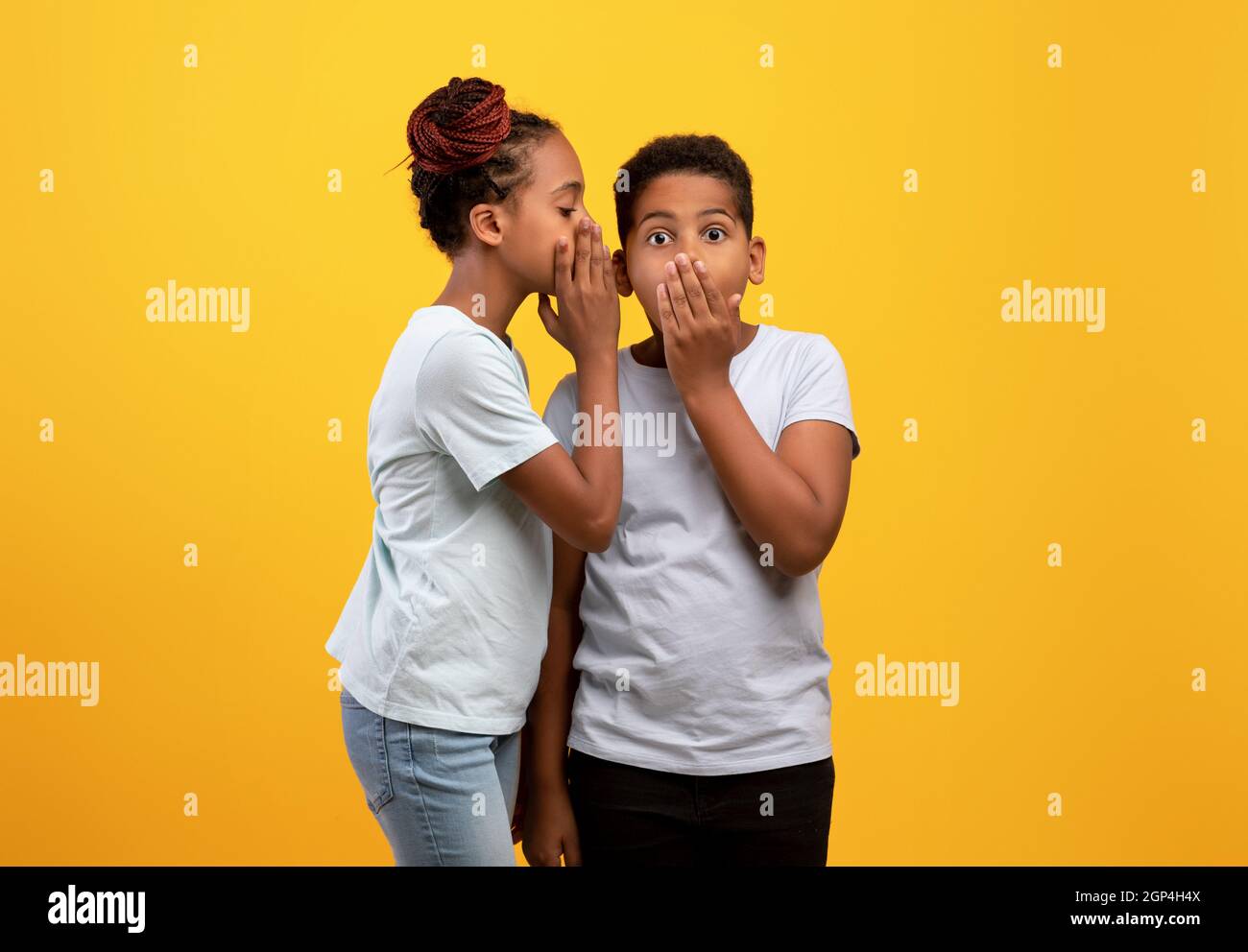 Black girl sister sharing secrets with her brother, african american teen girl whispering something to cute boy sibling, yellow studio background. Bro Stock Photo