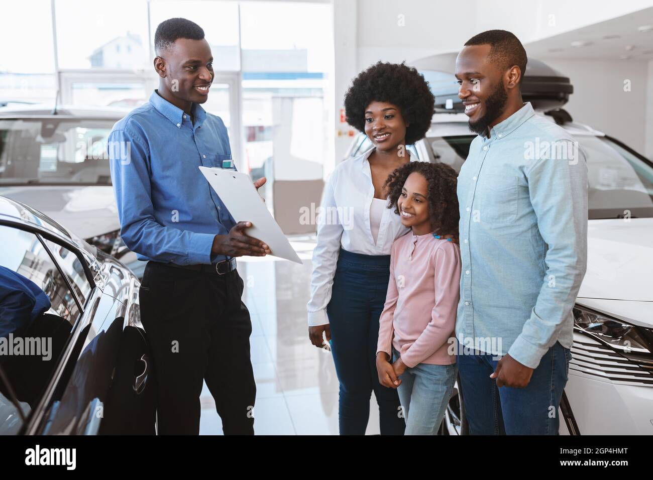 Positive black salesman offering Afro family to sign auto purchase or rental agreement at dealership. Cheerful young clients with cute kid buying new Stock Photo
