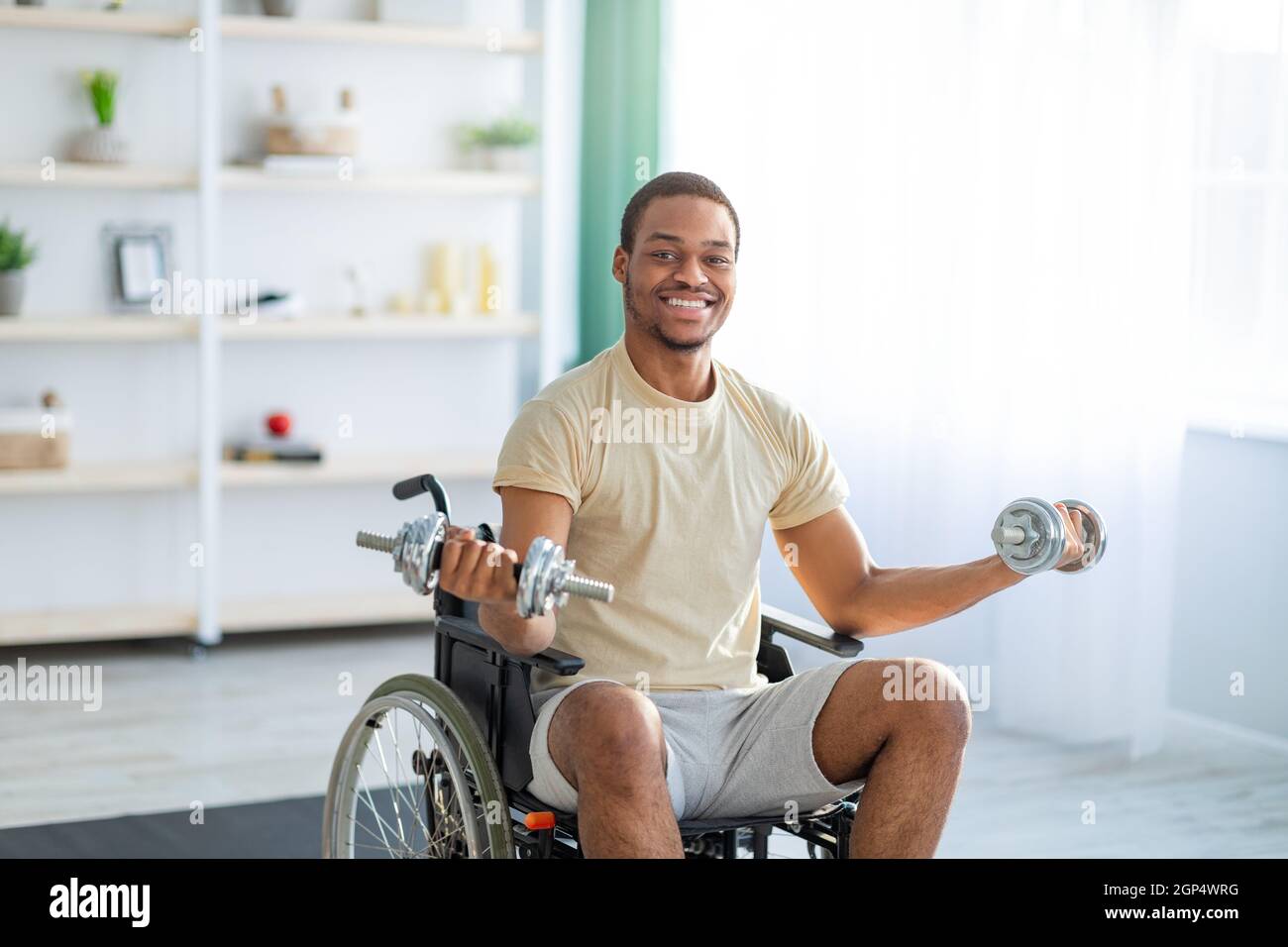 Physical activities for paraplegic people. Impaired black man in wheelchair making rehab exerises with dumbbells at home. Disabled young guy training Stock Photo