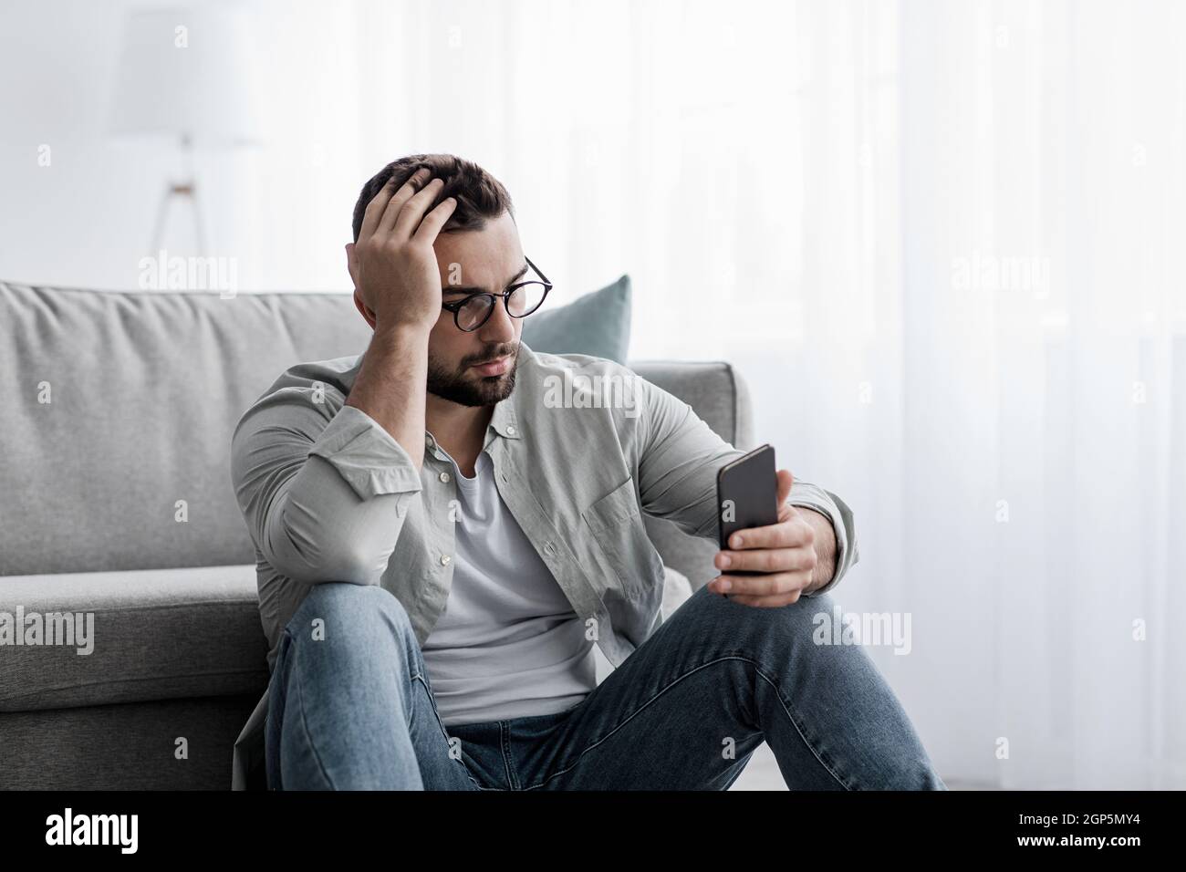 Relationship problems, facial expressions, bad news and messages, negative, stress and sad. Unhappy young handsome man in glasses sits on floor in liv Stock Photo