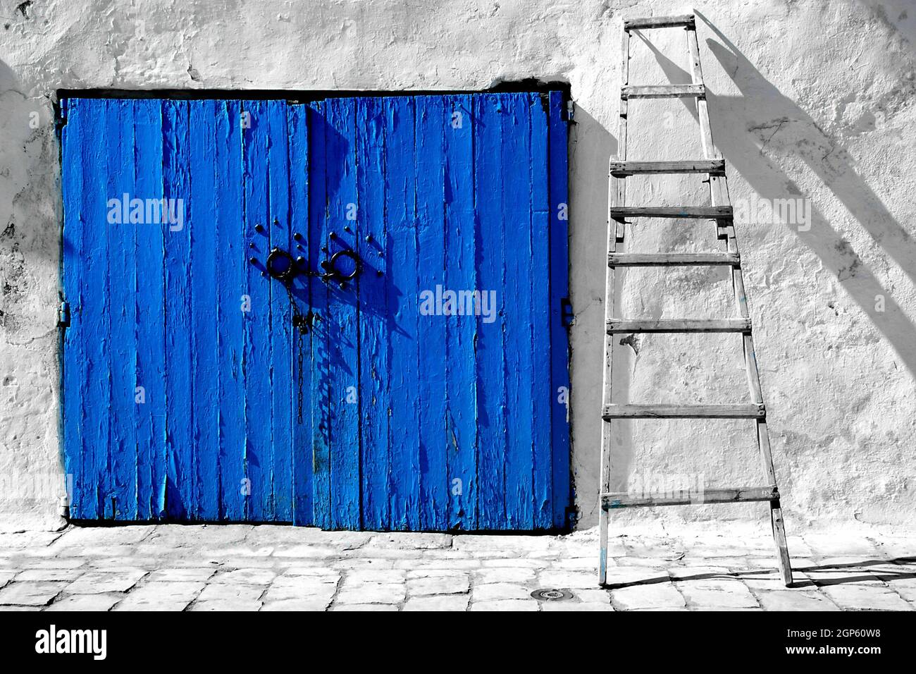 View of a blue wooden door with metal handles and ladder leaning on the wall next to it Stock Photo