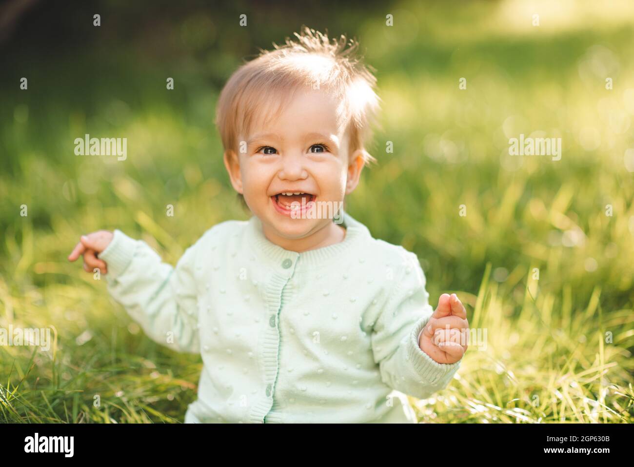 Cute laughing blonde baby girl 1 year old wear knit sweater top having fun sitting in green