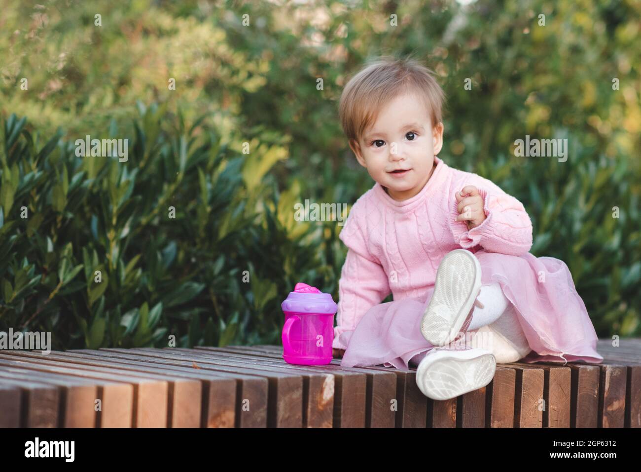 Cute funny baby girl 1 year old wear pink dress sitting with plastic bottle with water in park over green nature background close up. Looking at camer Stock Photo