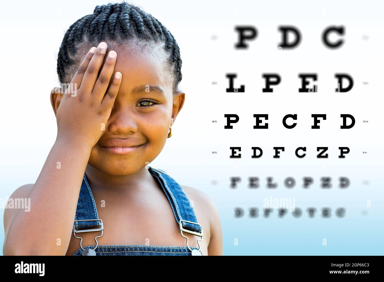 Close up Face shot of little African girl testing vision. Girl with braided hairstyle closing on eye with hand. Vision chart with block letters and fo Stock Photo