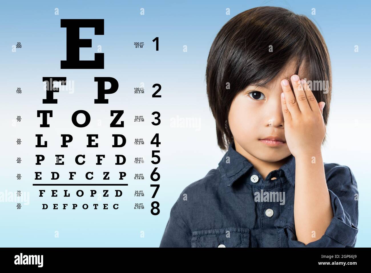 Close up portrait of little Asian boy reviewing eyesight. Kid closing one eye with hand and  alphabetical eye test chart in background. Stock Photo