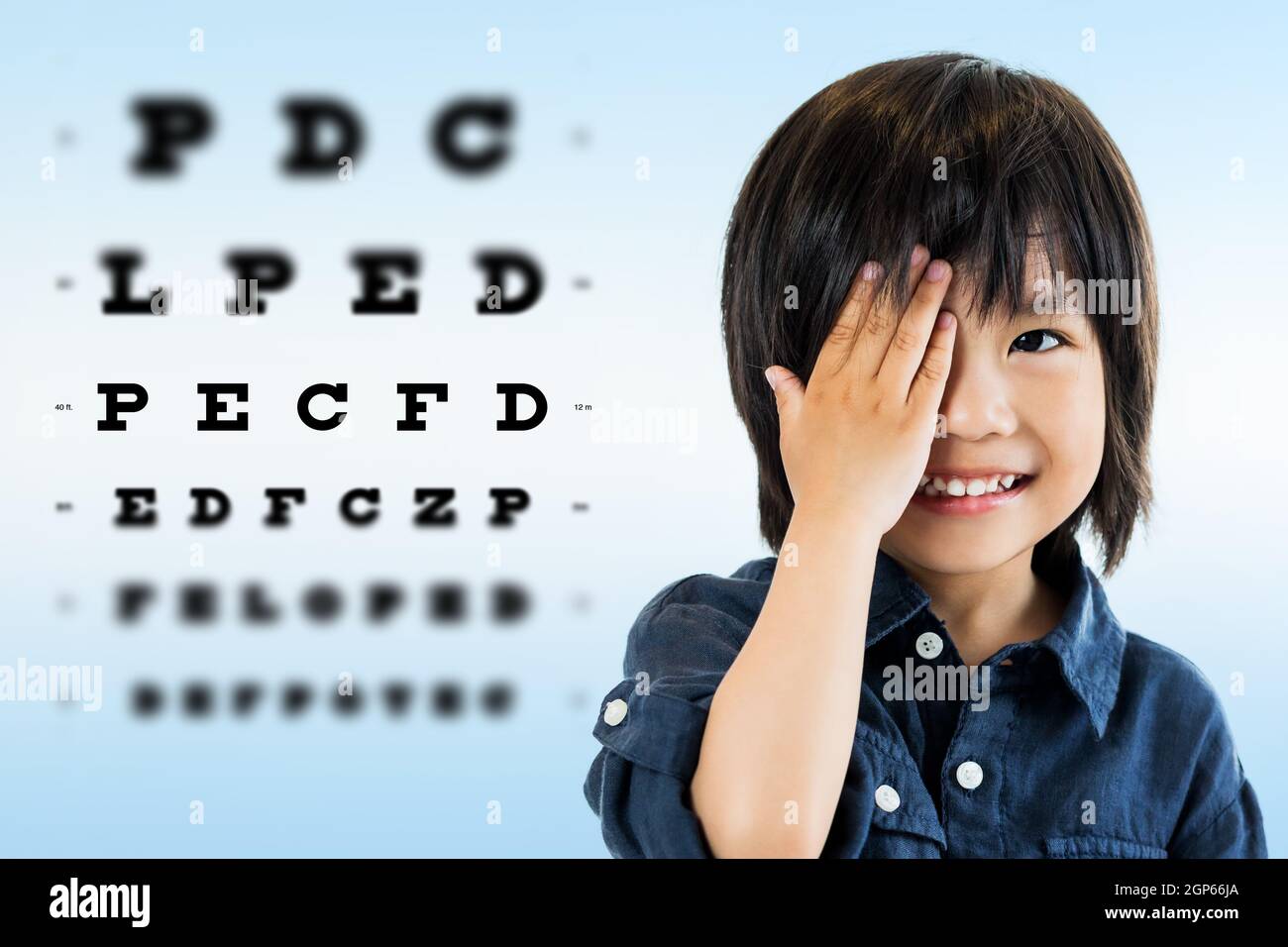 Close up portrait of cute little asian boy doing eye test.Kid closing one eye with hand against alphabetical out of focus test chart in background. Stock Photo