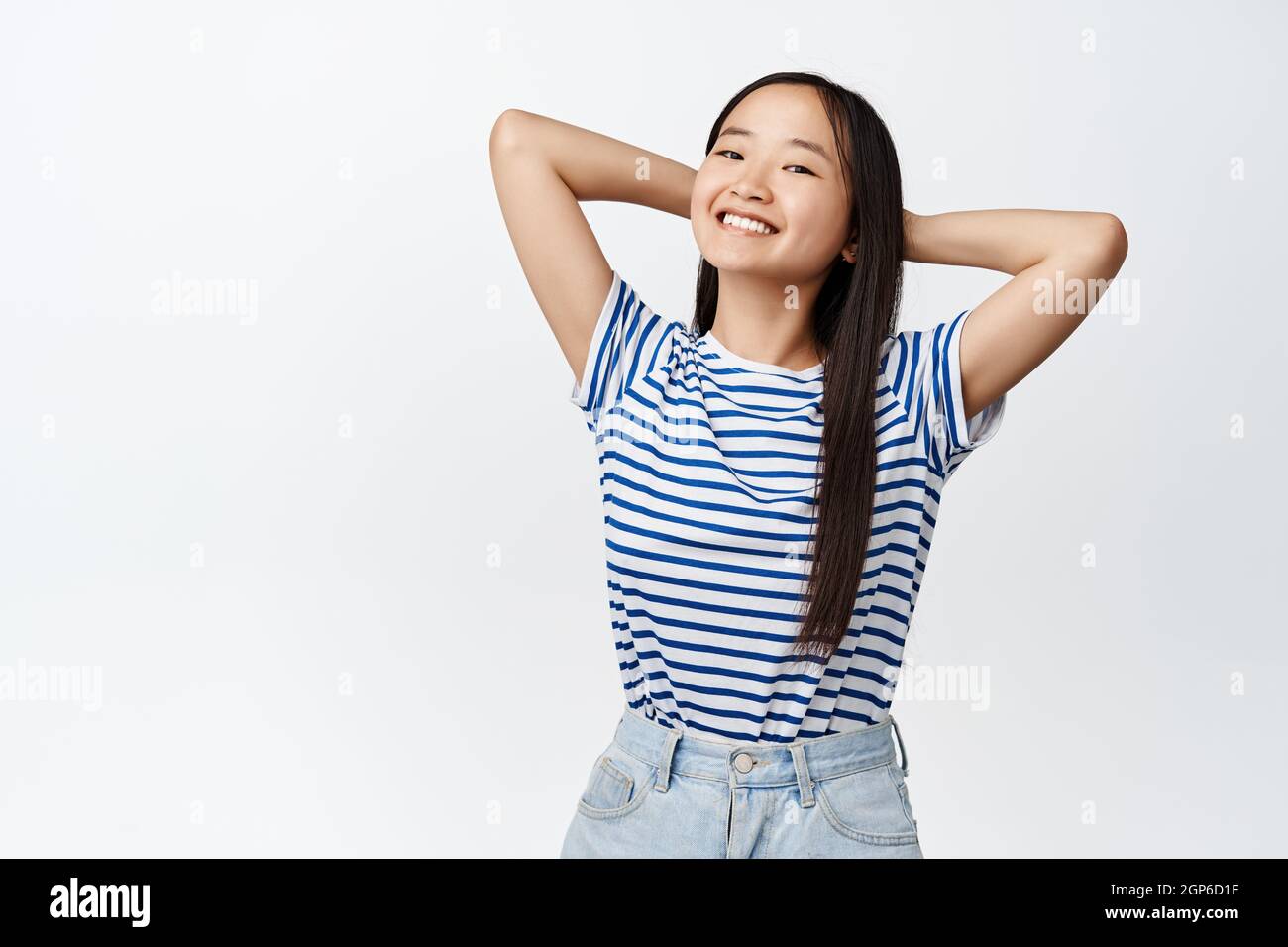 Portrait of brunette asian girl relaxing, holding hands behind head and watching something while resting, smiling satisfied, standing over white Stock Photo