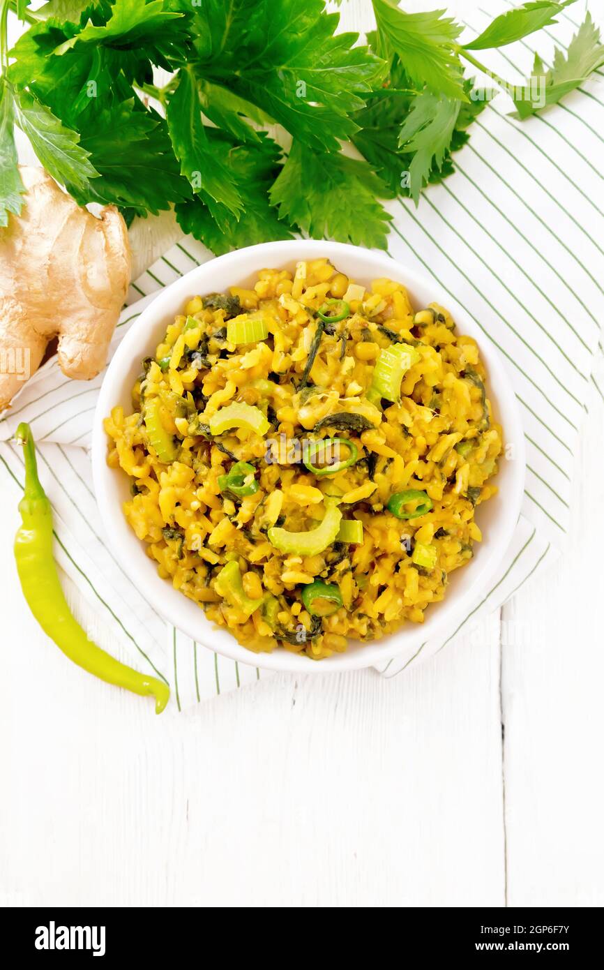 Indian national dish kichari made from mung bean, rice, stalk celery, spinach, hot pepper and spices in a bowl on a napkin, ginger on light wooden boa Stock Photo