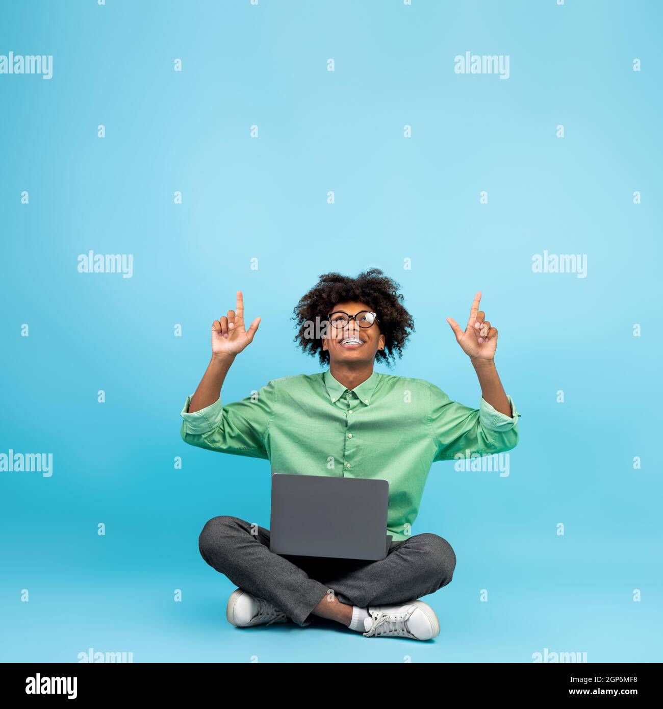 Check this. Black teen guy sitting with laptop and pointing up at free space above head over blue background, crop. Cheerful male student showing empt Stock Photo