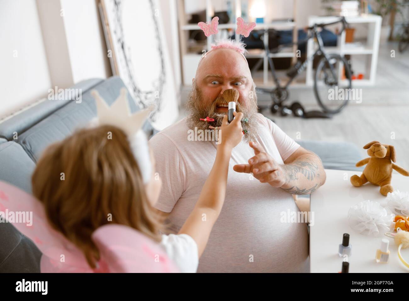 Daughter with princess crown applies powder onto nose of shocked father in living room Stock Photo