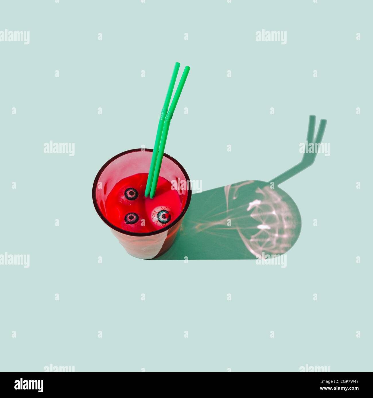 A glass filled with red drink, with eye balls in the liquid and two green straws. Pastel mint background. Spooky Halloween traditional celebration con Stock Photo