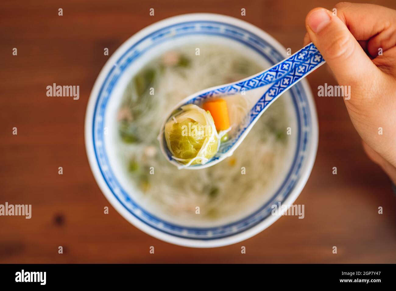 Top down view of a bowl of soup and a hand holding a spoon with cabbage and carrots and noodles. Stock Photo